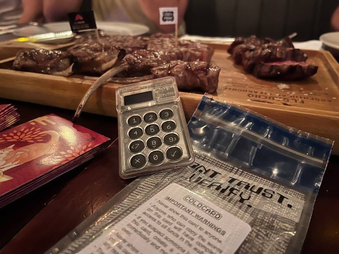 A humorous scene on a dinner table featuring grilled steaks, a mini calculator, two red Chinese New Year envelopes, and a tamper-evident Coldcard security wallet packaging labeled 'DON'T TRUST, VERIFY' with important warnings about sealed bags.