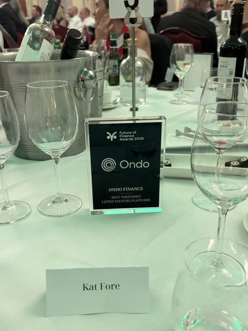 The image shows a table with several wine glasses, including some empty ones, placed on top of a white tablecloth. There are also multiple bottles of wine around the table. The main focus of the image is a plaque or sign that reads "KAT FORE" and "ONDO Finance". This suggests that the event may be related to the future of finance or tokenized equities platforms, as indicated by the post's content.