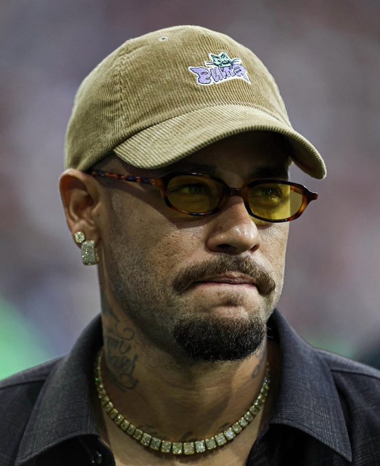 A close-up portrait of a man wearing a tan corduroy cap, yellow-tinted sunglasses, a diamond earring, and a diamond necklace, with visible neck tattoos.