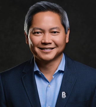 A professional headshot of an Asian man with graying hair wearing a dark blazer over a blue shirt, smiling at the camera against a dark background.