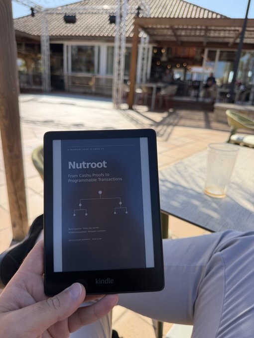 A hand holds a Kindle e-reader displaying the book 'Nutroot: From Cashu Proofs to Programmable Transactions' outdoors on a patio with a glass of water nearby.