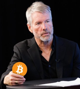 A man with gray hair and a beard in a dark blazer holds up a Bitcoin (₿) coin while sitting at a desk.