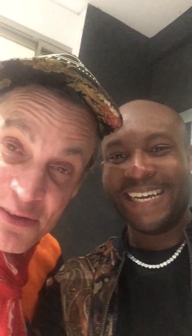 A selfie of two men smiling together indoors, one wearing a patterned head wrap and orange shirt, the other in a black shirt with a pearl necklace.