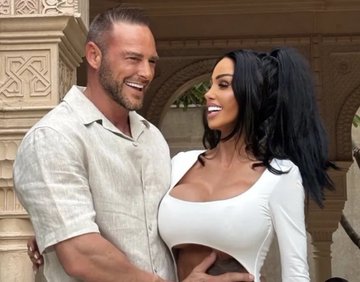 A muscular man in a short-sleeve button-up shirt stands smiling next to a woman in a white cut-out dress in a courtyard.