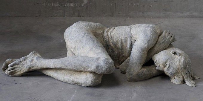 A weathered stone or plaster sculpture depicts a human figure curled in a fetal position on a gray floor, with the face resting against one hand.