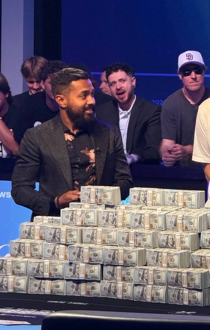 A bearded man sits behind a massive stack of bundled US dollar bills, surrounded by onlookers including someone in an SD Padres cap, appearing to celebrate a major poker or gambling win.