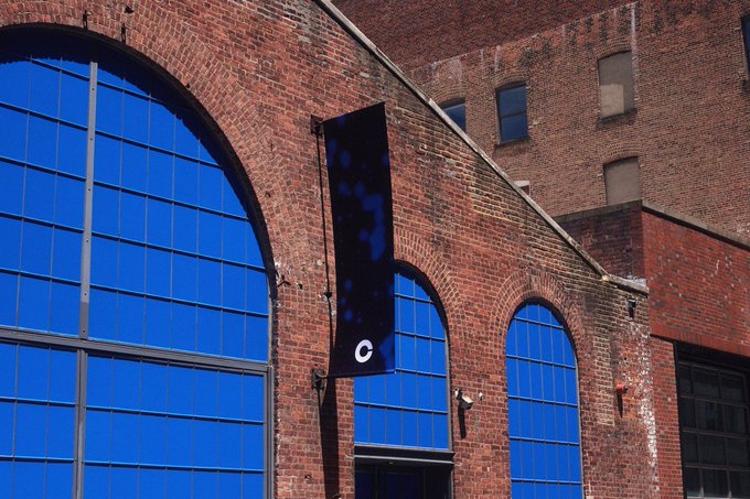 A brick building with large blue arched windows and a black vertical banner displaying a 'C' logo hanging on the facade.
