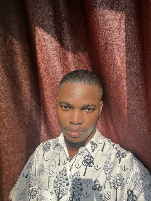 A young man wearing a white shirt with a black tree pattern poses for a portrait in front of a dark red curtain.