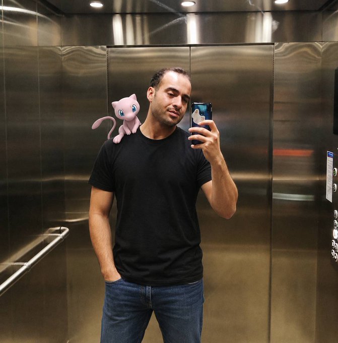 A man in a black t-shirt and jeans taking a selfie in an elevator with metallic walls, featuring a digital Mew Pokémon sticker edited onto his shoulder.
