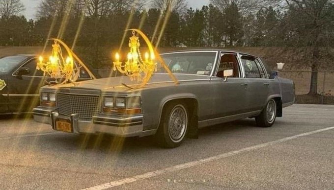 A classic Cadillac-style luxury sedan parked in a lot, humorously edited with glowing golden chandeliers mounted on its hood.