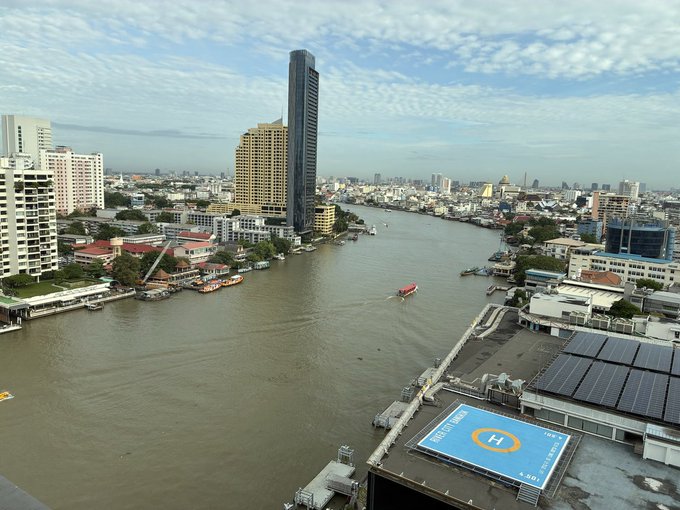 An aerial view of a city river with buildings on both banks, featuring a tall skyscraper and a helipad marked with 'H' on a rooftop.