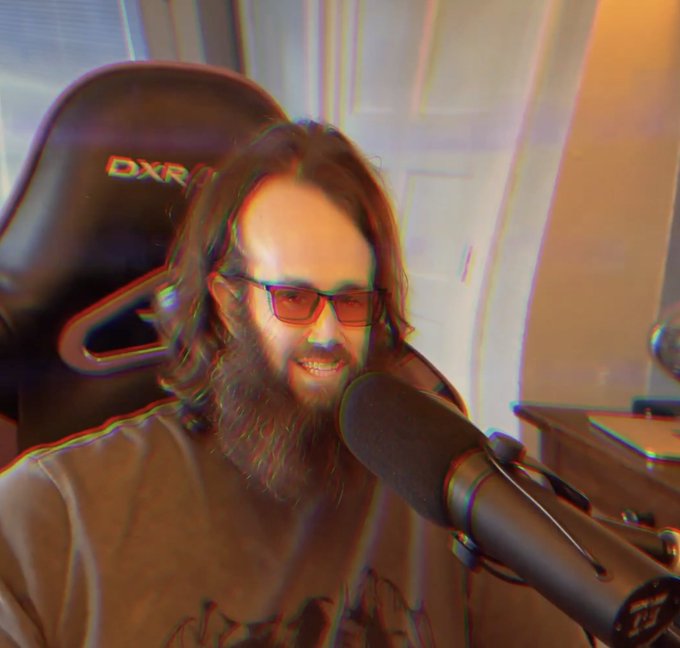 A man with long brown hair, a beard, and red-tinted glasses smiles while sitting in a DXRacer gaming chair, with a microphone visible in the foreground.