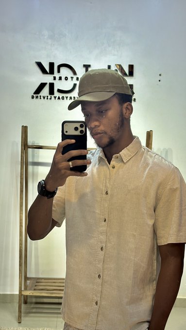 A young man in a beige shirt and cap takes a mirror selfie with his phone, standing in front of a wall that displays the partially visible "UNIQ..." logo (likely UNIQLO) in reverse reflection. The casual, friendly "hey" caption complements the laid-back, approachable vibe of the photo.