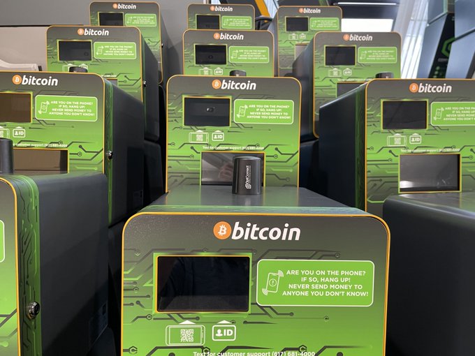 Several green Bitcoin ATMs are lined up, featuring screens and warning text about scams, including a message: 'ARE YOU ON THE PHONE? IF SO, HANG UP! NEVER SEND MONEY TO ANYONE YOU DON'T KNOW!'.