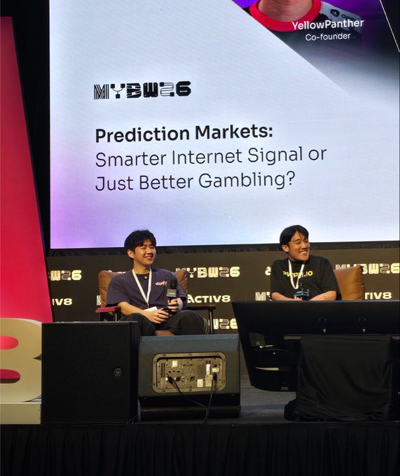 Two speakers sit on stage at MYBW6 (Malaysia Blockchain Week), discussing "Prediction Markets: Smarter Internet Signal or Just Better Gambling?" with YellowPanther, co-founder of Plume Network, introduced on the screen behind them.