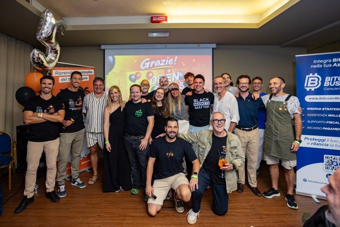 A group photo of around twenty people posing together at an event with a 'Grazie!' projection screen, balloons, and banners for 'BITS Business' and '4BLACK LIVES MATTER'.