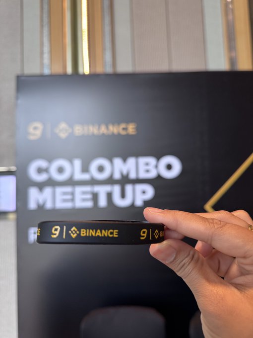 A hand holds up a black Binance wristband in front of a backdrop reading "BINANCE COLOMBO MEETUP," capturing a moment of community celebration that ties directly to the post's theme of unity, with a single branded band symbolizing the shared bond among Binance members marking the company's 9th anniversary.