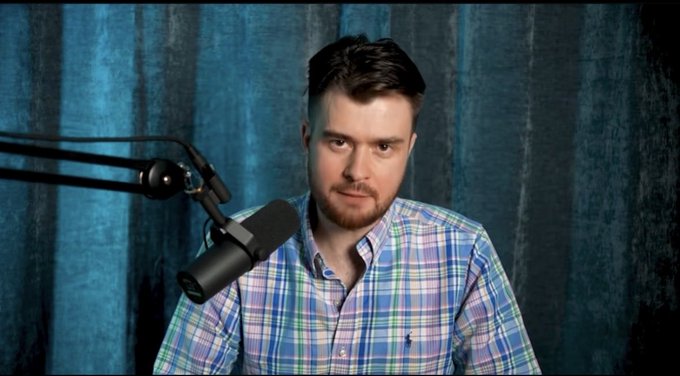 A man with brown hair and a beard, wearing a blue and green plaid shirt, sitting in front of a microphone against a blue curtain backdrop.