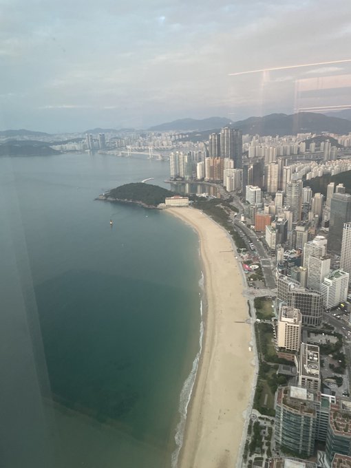 An aerial view of a coastal city with a curved sandy beach, high-rise buildings, and mountains in the distance.