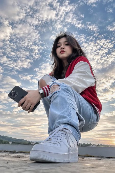 A young woman crouching on a rooftop holding a smartphone, wearing a red and white varsity jacket, ripped jeans, and white platform sneakers, photographed from a low angle against a cloudy sunset sky.