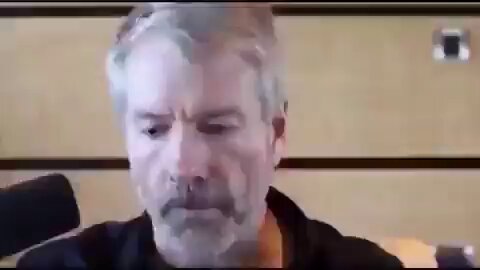 The image features an older man with gray hair wearing a black shirt, sitting at a desk or table while looking down at something. The background of the image shows a wooden wall, which could be part of a room or office setting. There is also a microphone visible in the scene, suggesting that this might be a recording session or a video call. The caption "It will turn out in the end That Saylor and everyone just needed a Monero" implies that there may have been a discussion about video games or possibly related to a gaming event.
