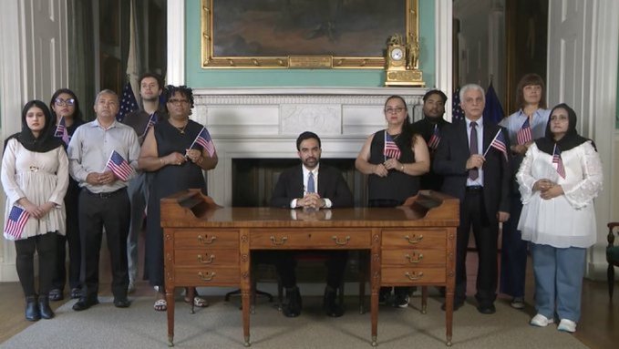 A group of people holding small American flags standing behind a man in a suit seated at a wooden desk in what appears to be an official government setting with a fireplace and painting in the background.