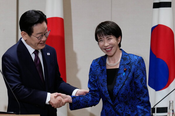 A man in a suit, wearing glasses, shakes hands with a smiling woman in a blue jacket. Flags of Japan and South Korea are visible in the background.