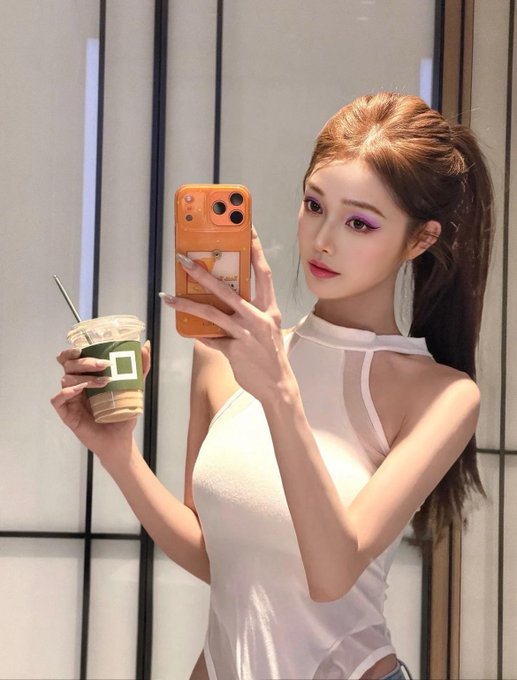 A young woman with purple eyeshadow taking a mirror selfie with an iPhone while holding an iced coffee drink.