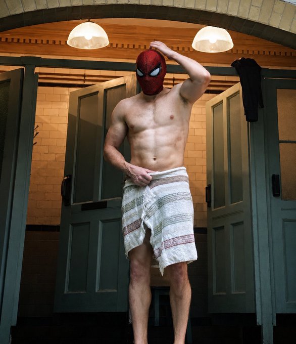 A shirtless, muscular man wearing only a Spider-Man mask and a striped towel stands in a doorway between bathroom stalls.