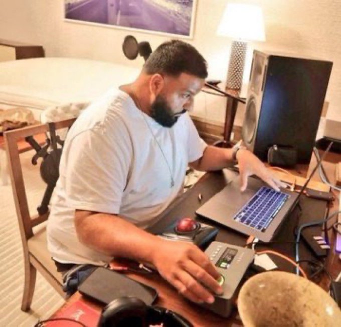 A bearded man in a gray t-shirt works at a desk using a laptop and an audio interface in what appears to be a hotel room.