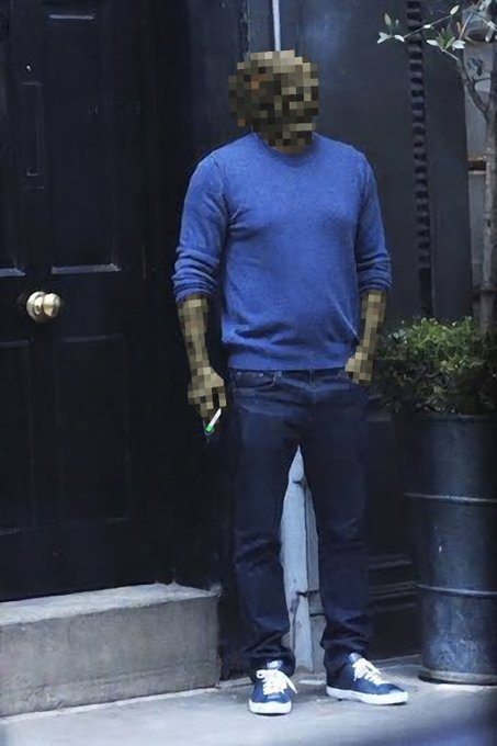 A person wearing a blue sweater, jeans, and blue sneakers stands against a dark door with a potted plant beside them; their face and hands are pixelated.