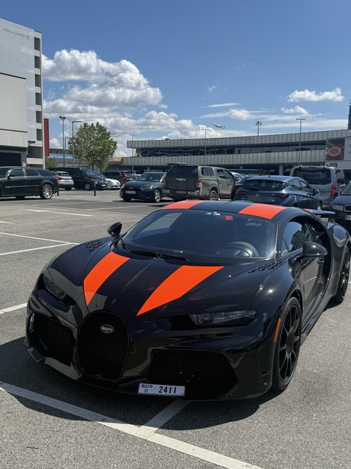 A black Bugatti Pur Sport sports car with orange racing stripes sits parked in a parking lot under a partly cloudy sky.