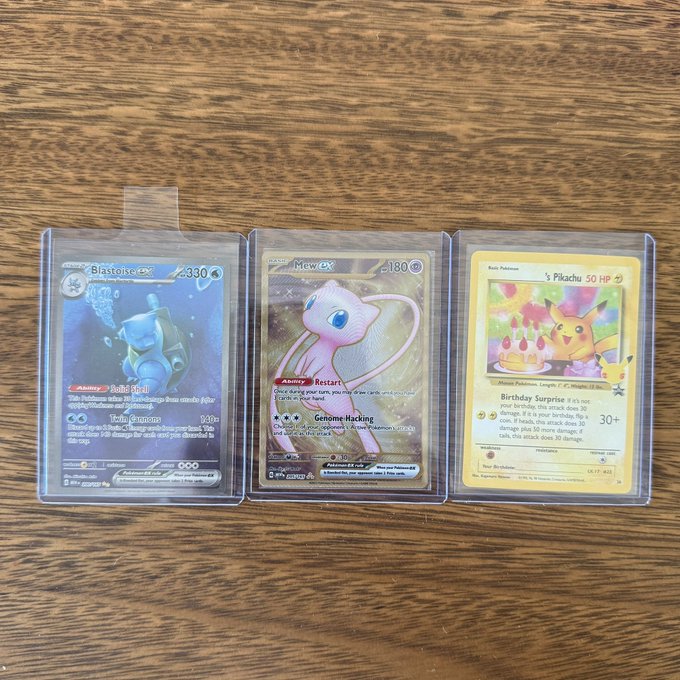 Three Pokémon trading cards are displayed on a wooden surface in protective sleeves: a Blastoise ex card, a Mew ex card, and a Pikachu birthday-themed card.