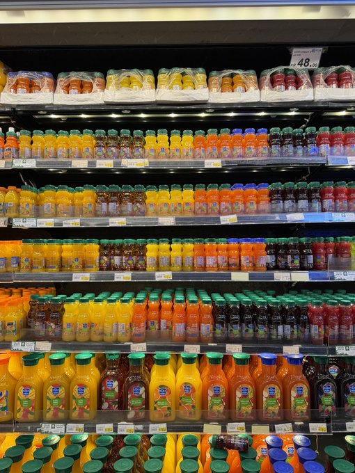 A well-stocked supermarket refrigerated aisle displaying rows of bottled juices and beverages in various colors, with packaged fruits on the top shelf.