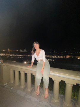 A woman in a white top and light green pants leaning on a balcony railing at night, with a cityscape visible in the background.