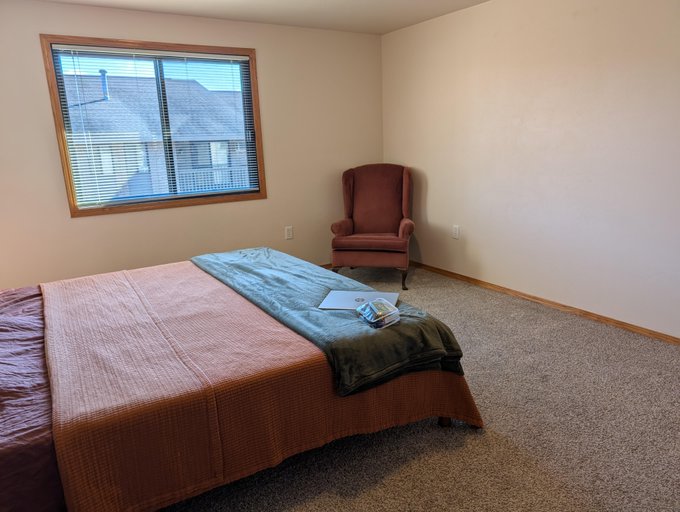 An empty bedroom contains a large bed with layered blankets, a maroon wingback chair in the corner, and a window with closed blinds.