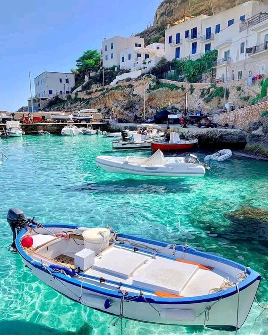A scenic photograph of a Mediterranean harbor with crystal-clear turquoise water, small white boats floating in the bay, and white buildings perched on a rocky cliff in the background.