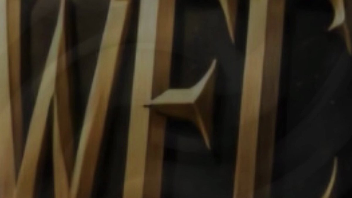 The image features a close-up view of a wooden object, possibly a clock or a decorative piece, with a gold design on its surface. The background appears to be black, which contrasts with the golden elements on the object. The focus is primarily on the intricate details and craftsmanship of the wooden item, while the surrounding environment seems to be intentionally blurred out for emphasis.
