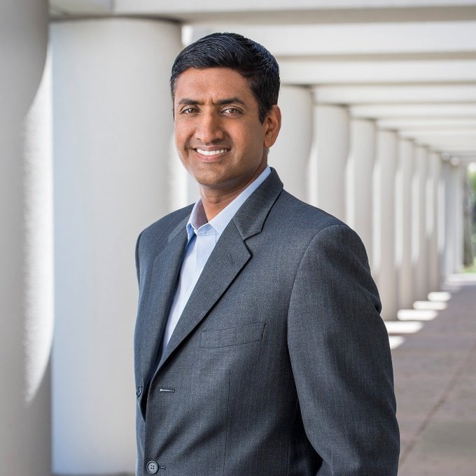 A smiling man in a gray suit and light blue shirt stands in front of a corridor lined with white columns. The image is unrelated to the accompanying post about Rep. Ro Khanna's disclosed trade of $HOOD stock.