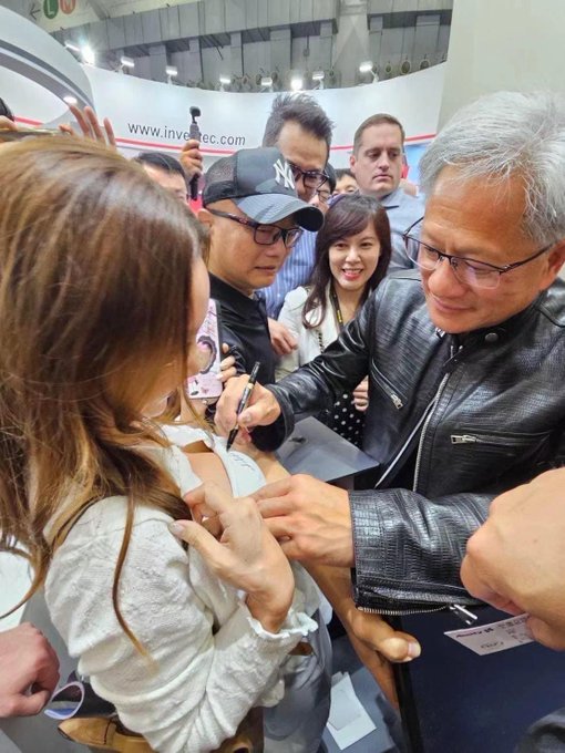 The image shows a man in a black leather jacket signing an autograph or item for a fan at what appears to be a convention, with a partially visible banner reading "www.investec.com" in the background, connecting to the post about the "$ai" stock target of 1 trillion in an investing context.