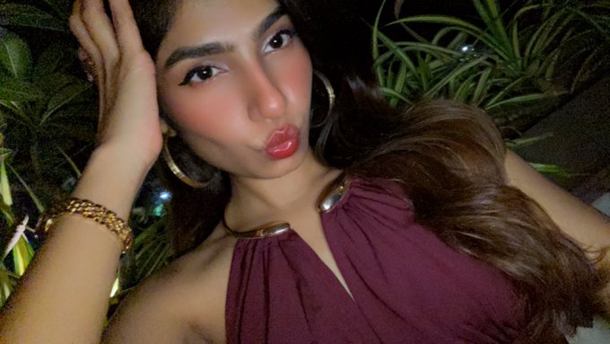 A selfie-style portrait of a young woman with long brown hair, gold hoop earrings, a gold watch, and a maroon halter top posing with red lipstick against a backdrop of green plants.