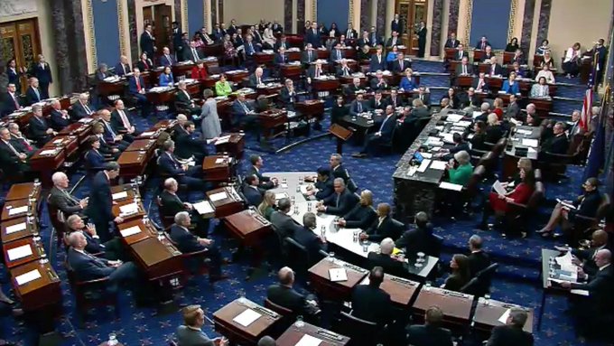 A wide overhead view of a crowded legislative chamber with senators seated at curved desks, with several individuals standing throughout the room.
