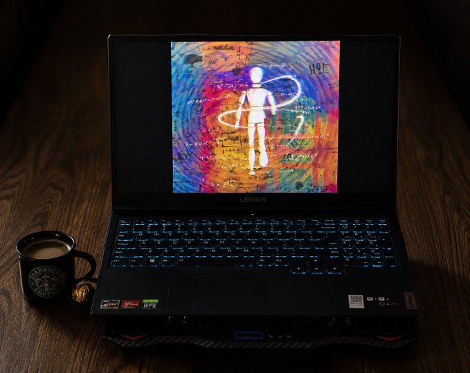 A Lenovo Legion gaming laptop with a backlit blue keyboard sits on a wooden table beside a black Starbucks mug; the screen displays a colorful abstract wallpaper with a glowing human figure.