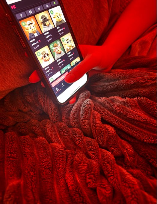 A close-up shot of a person in a red-lit environment using a smartphone app that displays a collection of cartoon-style avatar NFTs or digital characters, with the user's hand hovering over the screen.