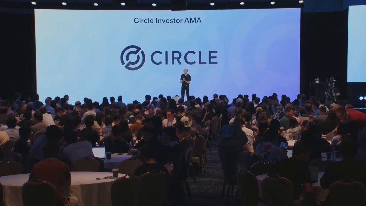 The image shows a large group of people gathered around a stage at a conference or event. A man is standing behind a podium, giving a speech or presentation. The audience appears engaged and attentive as they listen to him. The setting suggests that this could be an AI-powered event management system in action, where agents are responsible for various tasks such as finding clients, closing deals, and managing payments among others.