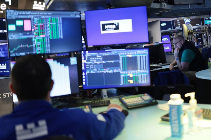 A trader in a blue NYSE jacket works at a desk filled with multiple monitors displaying financial charts and data, with an NYSE banner visible on one screen.