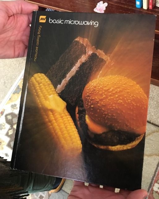 Hand holding a vintage 'basic microwaving' cookbook featuring a photo of a hamburger, corn on the cob, and a slice of cake on the cover.