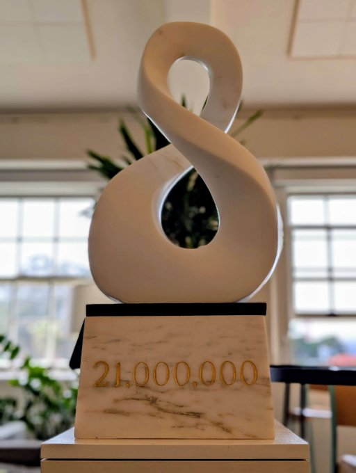 A white marble abstract sculpture in the shape of an infinity symbol or figure-eight, mounted on a rectangular base engraved with the number '21,000,000'.