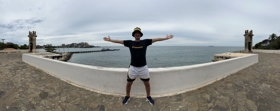 A panoramic photo of a man wearing a black Binance t-shirt and a yellow cap, standing with arms outstretched on a stone overlook by the sea.