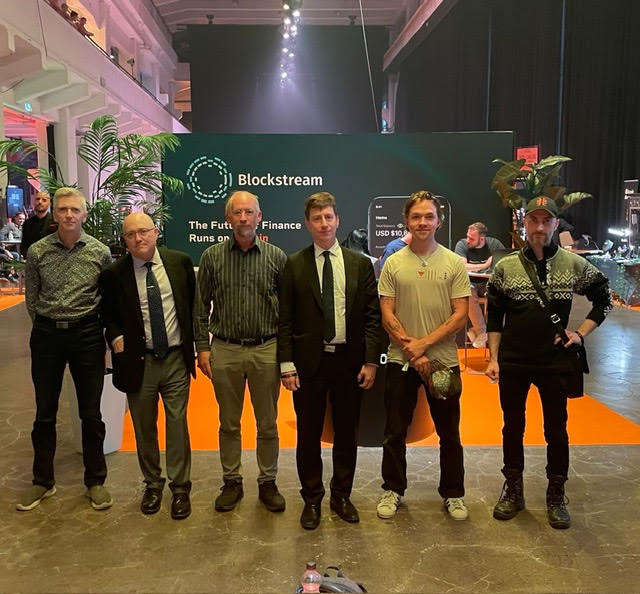Six men pose together in front of a green 'Blockstream' booth sign at a convention, with partial text reading 'The Fut... Financ... Runs on...' and 'USD $10...'.